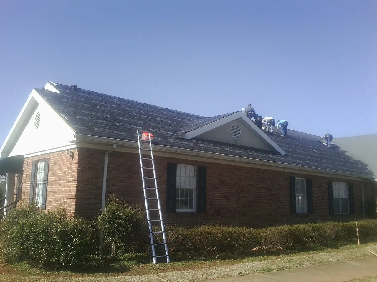 Crew tearing shingles off a brick ranch roof with a ladder set — Emergency Roof Repair work by our crew in Panora