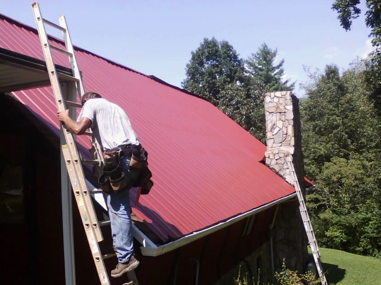 Roofer on a ladder beside a red metal roof and stone chimney — Soffit & Fascia Repair by Roofing Brothers in Panora
