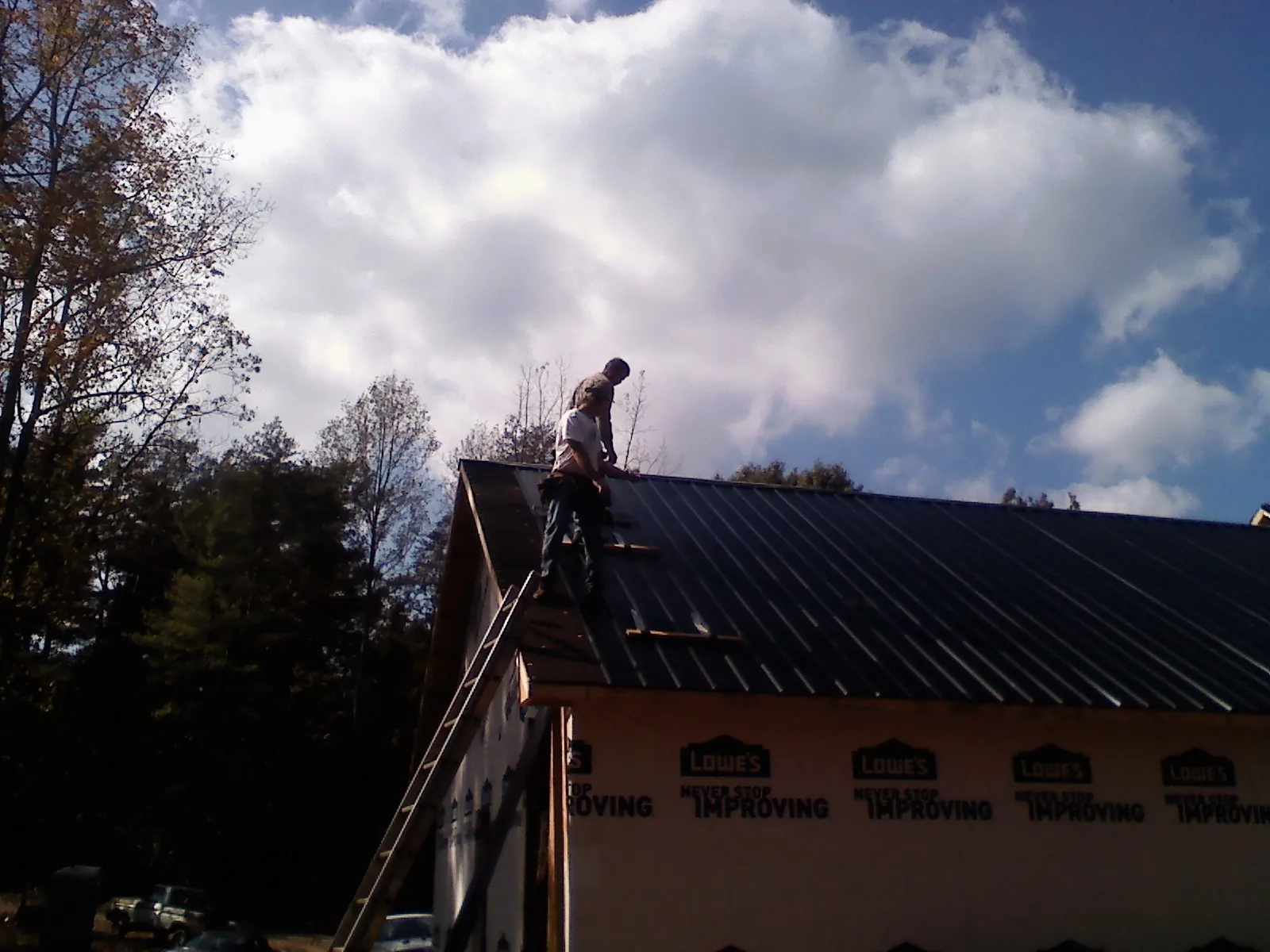 Roofer fastening dark metal panels high against a cloudy sky, from a Roof Inspection job in Panora